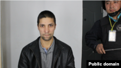 2018-жылдын февраль айында кармалган шинжаңдык Ильхам Исмаилдин жанында күч кызматкери турат. Булак: xinjiangpolicefiles.org