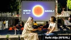 Dy gra bisedojnë, teksa në prapavijë shihet motoja e Dokufest-it të sivjetmë "Si të mbijetojmë".