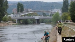 Punëtorë duke kositur bar buzë lumit Ibër, afër urës kryesore të qytetit të ndarë etnikisht, në Mitrovicë, më 9 shtator 2024.