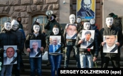 Революція гідності, Київ, 6 лютого 2014 року. Члени «Руху Гая Фокса» тримають портрети соратників президента Віктора Януковича під час акції «Похорон Партії регіонів» біля штабу партії у столиці України. Портрет Михайла Чечетова другий зліва
