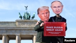 Участники акции протеста в Берлине в масках Дональда Трампа и Владимира Путина держат транспарант с лозунгом "Сделаем Россию снова великой"