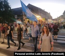 Lado Jovović, în timpul unuia dintre protestele din Novi Sad din 2023, poartă steagul orașului. Marija Vasić este și ea în fotografie.