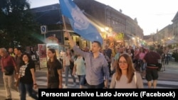 Lado Jovović, tokom jednog od protesta u Novom Sadu 2023. godine, nosi zastavu grada. Na fotografiji je i Marija Vasić.