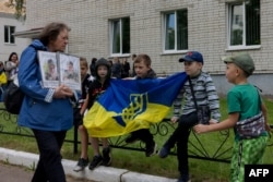 O femeie în mână cu fotografii ale prizonierilor trece pe lângă un grup de copii în timpul unui schimb de prizonieri de război într-o locație nedezvăluită din Ucraina, pe 12 iunie 2025.