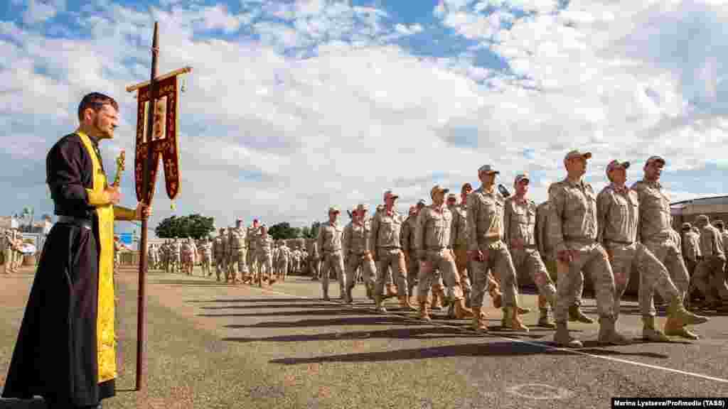 Ruske trupe marširaju pored pravoslavnog sveštenika u bazi Hmeimim u aprilu 2018. Vojnici nose ruske uniforme boje pijeska koje su izdate nekim vojnicima koji se nalaze u Siriji. Ben Dubow, viši saradnik u Centru za analizu evropske politike, rekao je za RSE da bi trenutni pregovori između Rusije i HTS-a&nbsp;mogli da dovedu do "znatno smanjenog ruskog prisustva" u bazama u Siriji u poređenju sa aktivnom fazom borbe tokom Assadove vladavine. &nbsp;